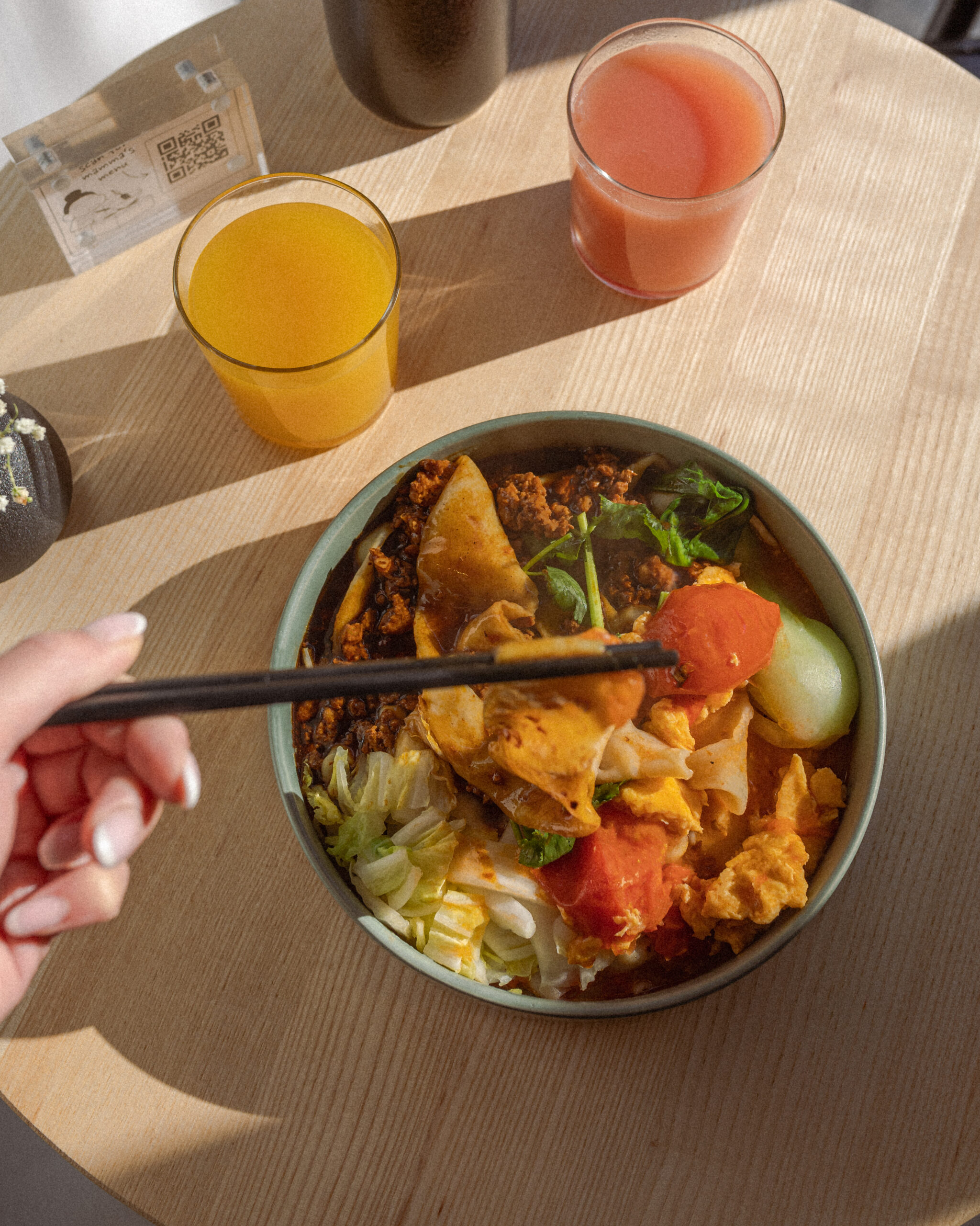 Eine Hand hält Stäbchen über einer asiatischen Nudelschüssel mit Hackfleisch, Tomaten und Gemüse, daneben Orangen- und Grapefruitsaft